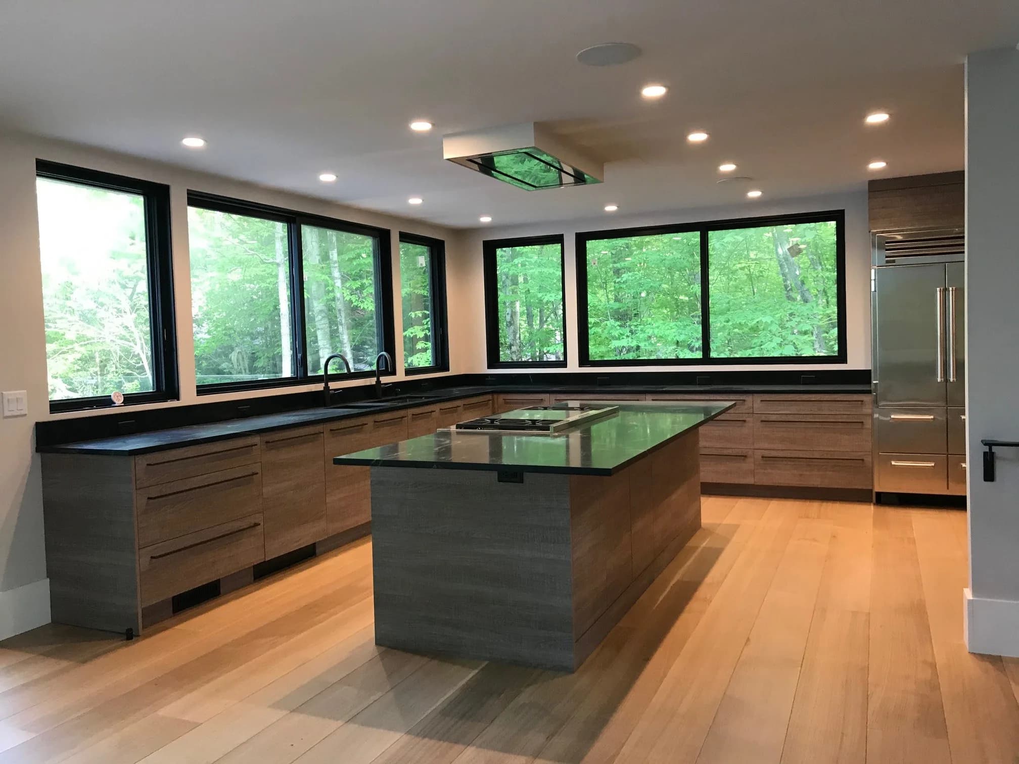 Kennebunk ME kitchen — wood-grain flat-panel cabinets, black stone island, large windows overlooking woods